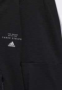 Svart atletisk klädsel med ribbad textur, med vit text "MÄRKET MED TRE STRIPOR" och Adidas-logotypen.