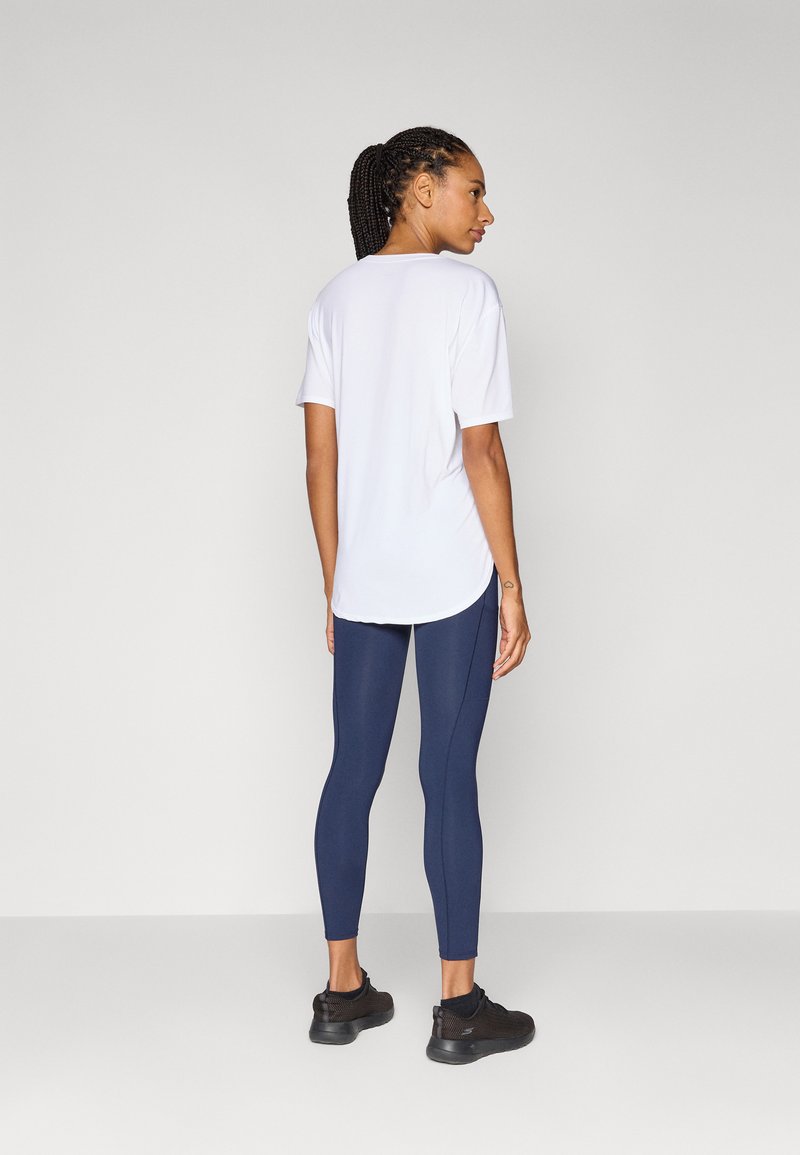 Eine Frau mit geflochtenen Haaren trägt ein weißes Shirt und marineblaue Leggings, steht seitlich und präsentiert sportliche Schuhe vor einem neutralen Hintergrund.