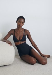Schwarzes Bralette und hochgeschnittene Panties, glattes Material, minimalistisches Design, taillierte Form, keine sichtbaren Muster, weiche Textur vor hellem Hintergrund.