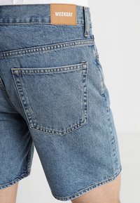 Pantalones cortos de mezclilla azul con un parche de cuero etiquetado "WEEKDAY", diseño de cinco bolsillos, dobladillo deshilachado y detalles de costura visibles.