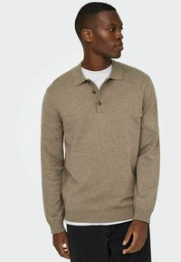 Polo de manga larga color marrón claro con cuello doblado y tapeta de tres botones. Textura suave, silueta ajustada, superpuesto sobre una camiseta blanca.