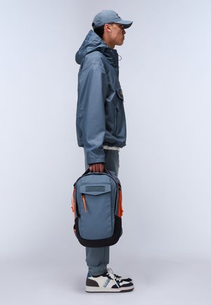 Grauer Rucksack mit schwarzen Akzenten und orangefarbenen Reißverschlüssen, rechteckige Form, Stoffstruktur, von einer Person in einem passenden grauen Outfit gehalten.
