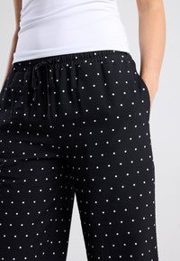 Black pajama pants with white polka dots, featuring a relaxed fit, elastic waistband, drawstring, and side pockets. Soft fabric texture.