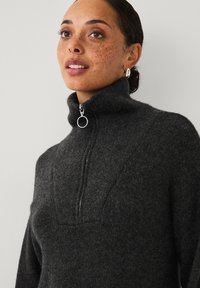 Kvinde med fregner iført en mørkegrå lynlås-sweater med høj krave og sølvfarvede hoopsøreringe, der ser let opad mod en lys baggrund.