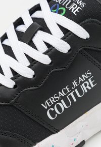 Schwarzer Sneaker mit weißen Schnürsenkeln und der Aufschrift "VERSACE JEANS COUTURE". Verfügt über ein strukturiertes Obermaterial und eine gesprenkelte Gummisohle.