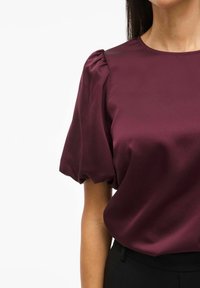 Blusa in satin bordeaux con scollo rotondo, maniche corte a palloncino e texture morbida. Abbinata a pantaloni neri, enfatizza la lucentezza del tessuto e la forma.