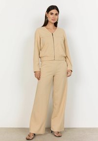 Beige svetr na zip s dvěma předními kapsami a odpovídajícími širokými kalhotami. Hladký materiál s volným střihem a elastickým pasem.