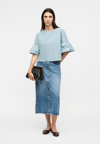 Γυναίκα που φοράει ανοιχτό μπλε μπλούζα με μανίκια με βολάν, μια τζιν midi φούστα με κουμπιά, κρατώντας ένα μαύρο τσαντάκι και καφέ επίπεδα παπούτσια.
