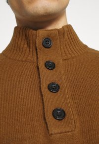 Bruine geribbelde gebreide trui met een hoge kraag en drie zwarte knopen langs de knoopsluiting. Zachte textuur en aansluitend ontwerp.