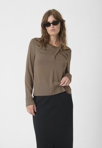 Haut marron à manches longues avec un col drapé, comportant une poche latérale. Porté avec une jupe noire, texture lisse et coupe décontractée.