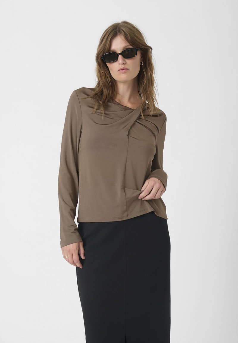 Brown long-sleeve top with a draped collar, featuring a side pocket. Worn with a black skirt, smooth texture, and relaxed fit.