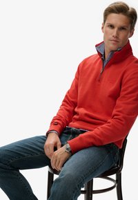 Sudadera roja con cremallera y cuello alto, combinada con una camisa de rayas azul clara, y unos vaqueros azules. En la muñeca del modelo se puede ver un reloj.