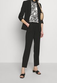 Černý blazer přes bílou košili s květinovým vzorem, doplněný černými kalhotami a plochými sandály. Materiály vypadají hladce a mají strukturovaný střih.
