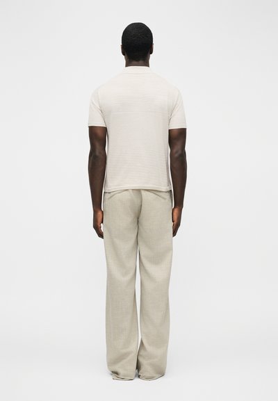 Άνδρας που στέκεται γυρισμένος προς τα πίσω, φορώντας κοντομάνικο μπεζ πουκάμισο και χαλαρό ανοιχτό μπεζ παντελόνι, μπροστά σε απλό φόντο.