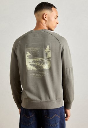 Homme portant un sweat à capuche vert olive avec un croquis de la nature et un texte sur la préservation du patrimoine naturel, associé à un jean bleu.