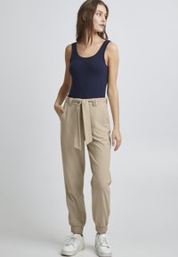 Beige lässige Hose mit elastischen Bündchen, tailliertem Bund und Seitentaschen. Kombiniert mit einem navyblauen ärmellosen Oberteil und weißen Sneakern.