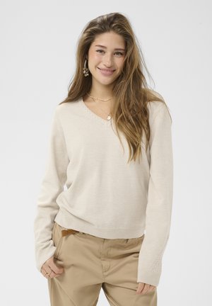 Jonge vrouw met lang bruin haar draagt een beige trui en lichtbruine broek, glimlacht met één hand in haar zak tegen een effen achtergrond.