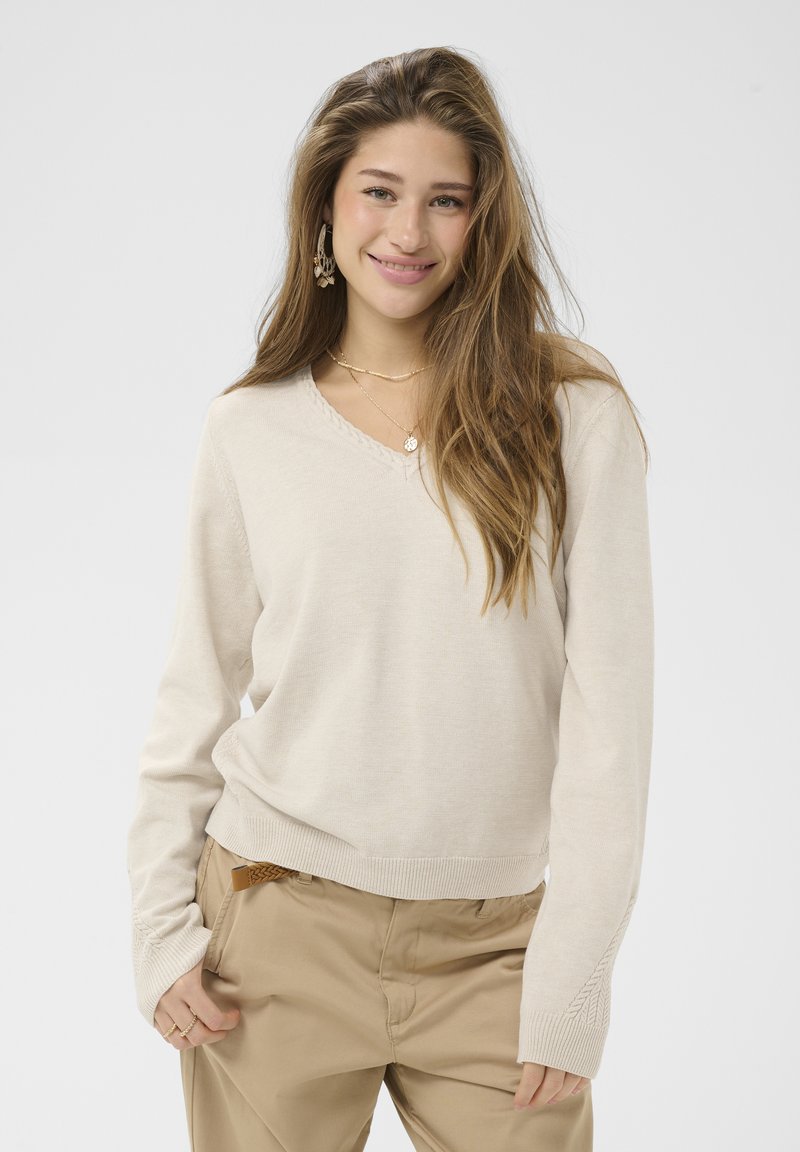 Jeune femme aux longs cheveux bruns porte un pull beige et un pantalon beige, souriante, une main dans la poche, sur un fond uni.