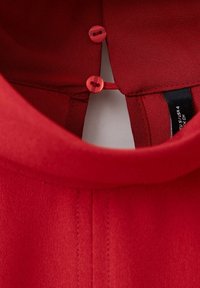 Gros plan d'un col de vêtement rouge avec deux petits boutons rouges retenus par une boucle, et une étiquette de taille noire partiellement visible à l'intérieur.