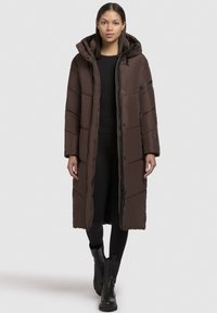 Palton lung umplut, de culoare maro, cu glugă, având închidere cu fermoar și capse, cusături verticale și un design elegant. Purtați peste un top și pantaloni negri.