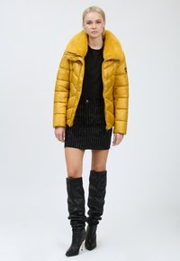 Gul quiltet pufferjakke med en bred faux fur-krave, parret med en sort nederdel med lodrette hvide striber og knæhøje sorte støvler.