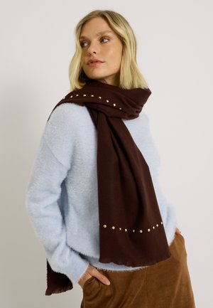Bufanda marrón hecha de un tejido suave, con tachuelas doradas dispuestas en una línea a lo largo de un borde. La mujer lleva un suéter azul claro y pantalones marrones.