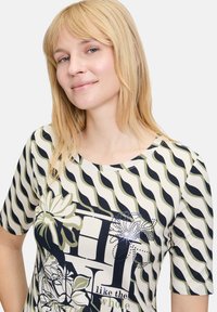 Short-sleeve t-shirt in cream with navy and olive wavy stripes, floral patterns, and graphic text elements. Soft fabric with a relaxed fit.