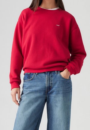Kvinde iført rød Levi's sweatshirt og blå jeans, med den ene hånd i lommen, foran en ensfarvet lys grå baggrund.
