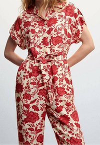 Mujer vistiendo un mono con botones de manga corta con estampados florales rojos y un cinturón atado a la cintura sobre un fondo crema.