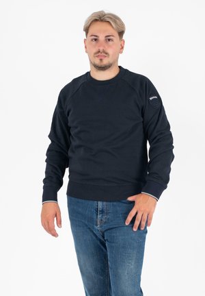 Felpa di cotone blu navy con maniche raglan e polsini a costine. Presenta un collo rotondo e una patch con logo sulla manica sinistra. Indossata con jeans di denim.