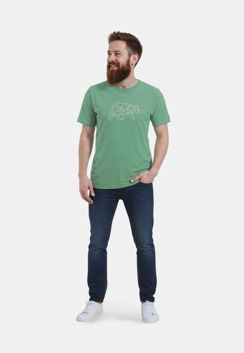 Grünes Baumwoll-T-Shirt mit einem weißen geometrischen Bärenmotiv auf der Vorderseite, kombiniert mit dunkelblauen Jeans und weißen Sneakern.