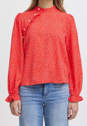 Blouse rouge à manches longues à pois blancs, boutons en perle et volants près de l'épaule et des poignets, associée à un jean bleu clair.