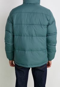 Grön puffjacka med hög hals, sydda paneler, elastiska ärmslut och en slät textur. Bärs över denimjeans.