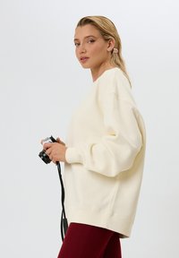 Felpa color crema con una vestibilità morbida, polsini e orlo a costine, abbinata a leggings bordeaux a coste, e una macchina fotografica in mano.