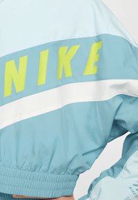 Geacă de vânt albastru deschis cu logo-ul „NIKE” galben neon, dungă orizontală albă, manșete elastice și textură netedă.