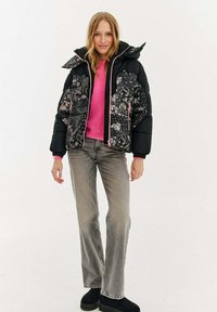 Kobieta ubrana w czarną puchową kurtkę w kwiatowy wzór, różowy sweter, szare dżinsy i czarne buty, stojąca na białym tle.