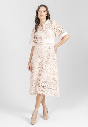 Kobieta stojąca w delikatnej różowej, koronkowej sukience midi z krótkimi rękawami, kołnierzykiem, z przodu z guzikami oraz z dopasowanym paskiem, ubrana w beżowe szpilki.