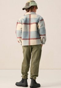 Veste en plaid en laine dans des tons bleus, crème et rouges, avec un col et des poignets à boutons, associée à un pantalon vert olive à taille élastique et des bottes noires.