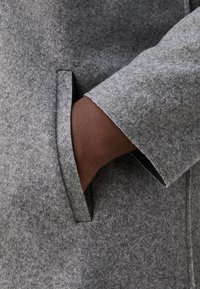 Szary wełniany płaszcz z detalami przy kieszeniach. Tkanina jest teksturowana i miękka, z dopasowanym krojem i wyraźnymi liniami.