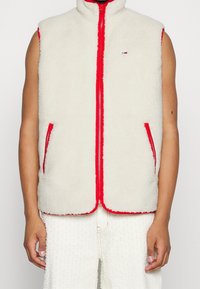 Fleecová vesta v krémové barvě s červeným zipem a lemováním. Má dvě boční kapsy. Bez rukávů s vysokým límcem, měkká textura.
