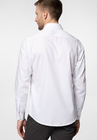 Biała koszula z długim rękawem o gładkiej fakturze, posiadająca klasyczny kołnierzyk oraz zaokrąglony dół. Widok z tyłu pokazuje smukły krój.