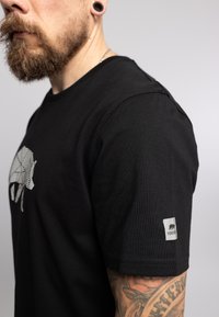 Camiseta de algodón negra con un gráfico de elefante a rayas en el frente y una pequeña etiqueta gris en la manga derecha. Diseño de manga corta, ajuste regular.