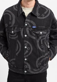 Una persona indossa una giacca di denim nera con un grande motivo logo sottile e bottoni bianchi, abbinata a una camicia di colore chiaro sotto.