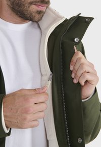 Dunkelgrüne Jacke mit einem weißen Fleece-Futter, Reißverschluss und Druckknopfverschlüssen. Die innere Schicht ist weich und sichtbar, mit kontrastierenden Texturen.