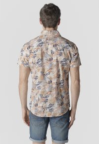 Hombre con camisa de manga corta beige con patrones de hojas naranjas y oscuras y pantalones cortos de mezclilla remangados, de espaldas sobre un fondo liso.
