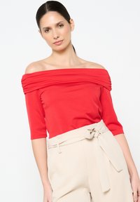 Top rosso a spalle scoperte con dettagli plissettati, maniche corte, abbinato a pantaloni a vita alta di un beige chiaro dotati di una cintura annodata.