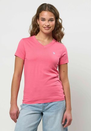 Tânără cu păr castaniu ondulat, purtând un tricou roz cu decolteu în V, mulat, și blugi deschiși la culoare, stând în fața unui fundal simplu.