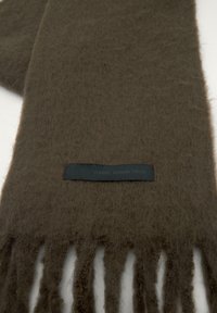 Sötét olívazöld gyapjú sál, puha, bolyhos textúrával, rojtos szélekkel és a "CLASSIC. MODERN. YOURS." felirattal.