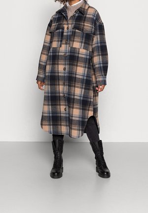 Vrouw die een lange geruite jas met knopen en grote borstzakken draagt, zwarte broek en zwarte veterlaarzen, staand tegen een effen achtergrond.