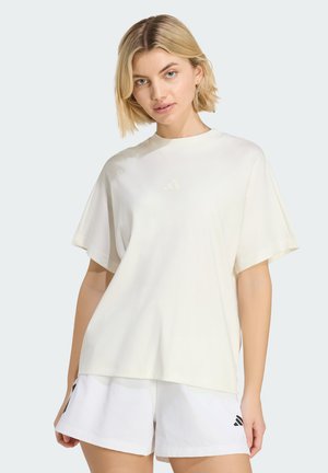 Lyshåret kvinde iført en løs hvid T-shirt og hvide shorts med sorte Adidas-logoer, stående mod en lys baggrund.